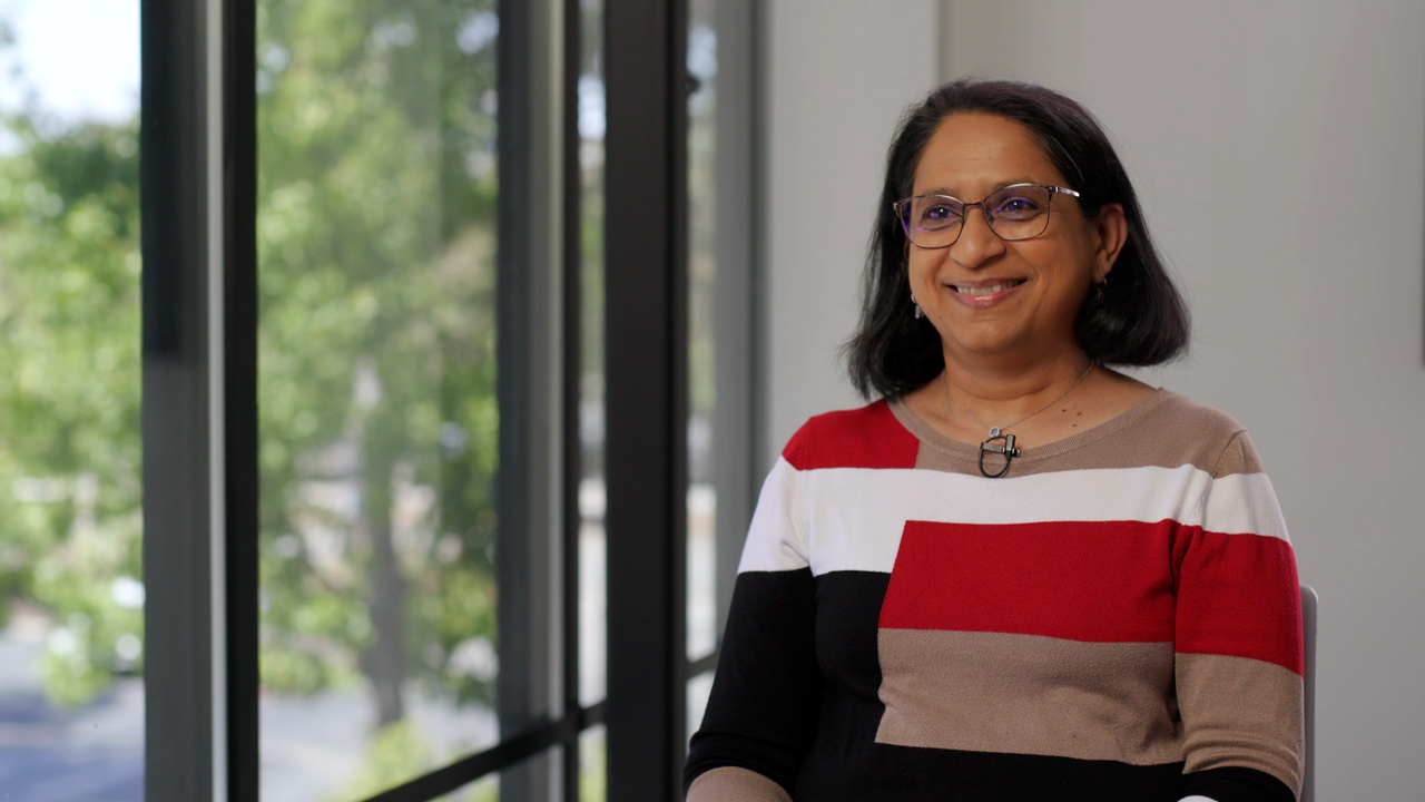 Woman in red, white, black and tan sweating sitting next to a window smiling.