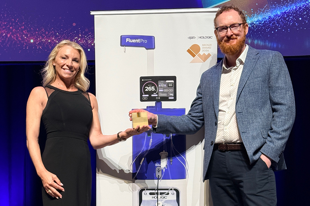 A man presents a bronze award trophy to a woman on stage beside a Hologic FluentPro banner.