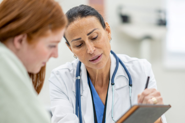 Woman speaks to provider wearing scrubs and holding a clipboard in exam room setting.